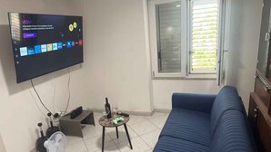 En stue med et vægmonteret fladskærms-tv, der viser en streamingtjenestes brugerflade. Der er en blå sofa, et lille bord med en flaske og et glas, og en ventilator på gulvet.