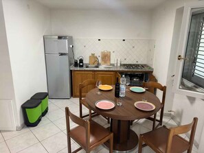 A compact kitchen with a refrigerator, sink, and stove. There is a dining table with chairs and plates. - Place in the Green | Apartment in Cala Gonone (Cala Gonone)