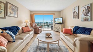 A living room with two beige sofas, a coffee table, a TV, and artwork on the walls. - New! Beachfront 2BR, Stunning Gulf Views, Indoor/Outdoor Pools, Hot Tubs (Gulf Shores)