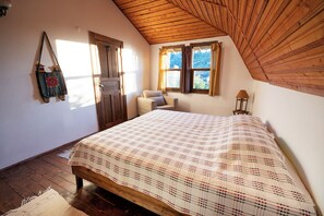 A bedroom with a wooden ceiling, a bed with a plaid bedspread, a chair, a small table with a lamp, and a door with curtains. - VİTA CAMPING (Konyaalti)