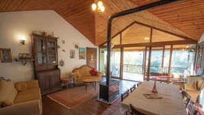 A cozy living room with wooden floors, a fireplace, a sofa, a dining table, and a large window offering a view of the outdoors.