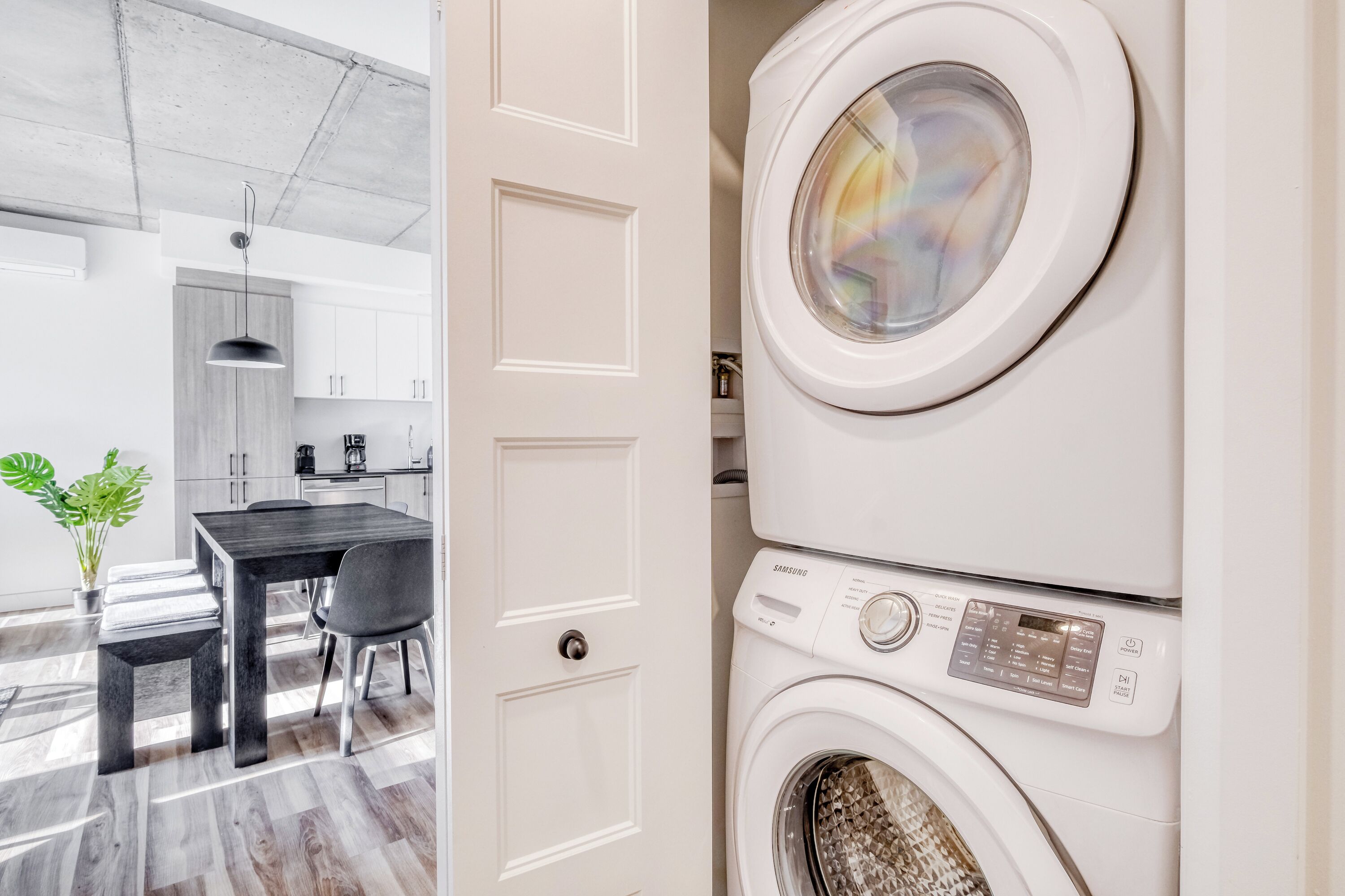 Une buanderie avec des lave-linge et des sèche-linge superposés, un coin repas avec une table et des chaises, et une plante en pot.