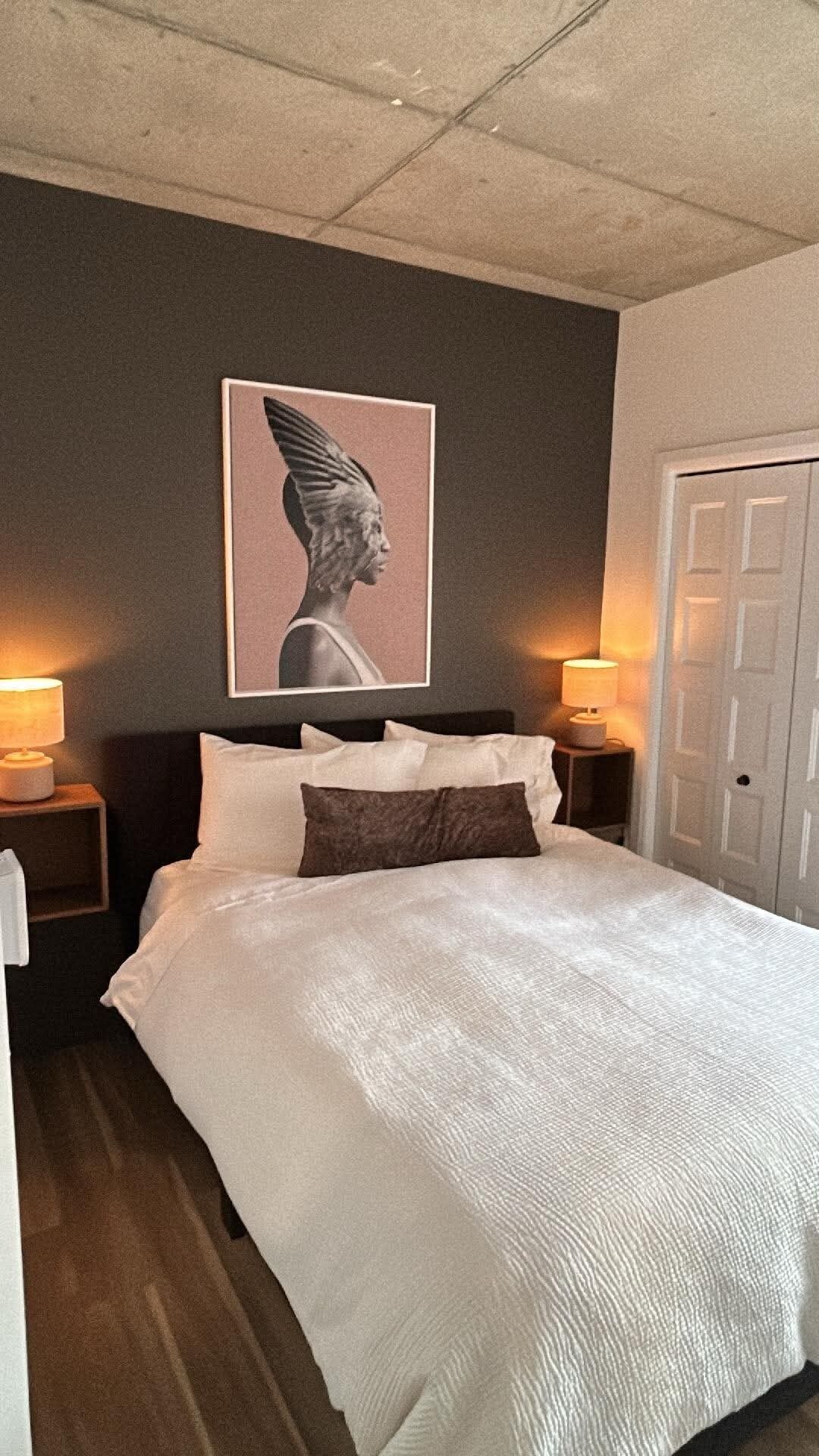 A bedroom with a bed, two bedside lamps, a door, and a wall-mounted artwork.