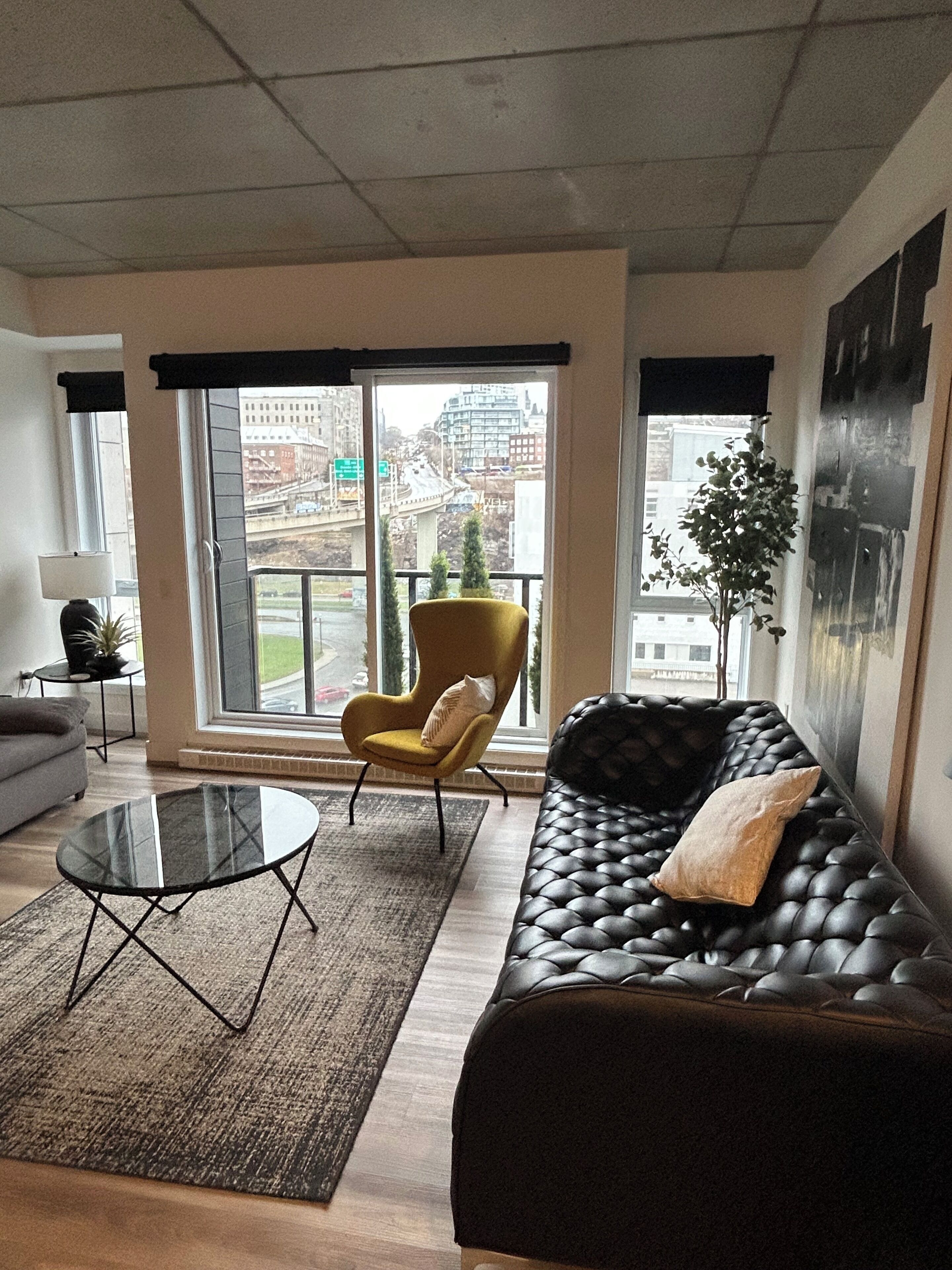A modern living room with a sofa, armchair, glass coffee table, and a view of the city through large windows.