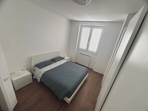 A bedroom with a bed, a nightstand, a window with blinds, and a radiator. - Quiet apartment in serviced area 10 minutes' drive from Milan. (Bettola-Zeloforamagno)
