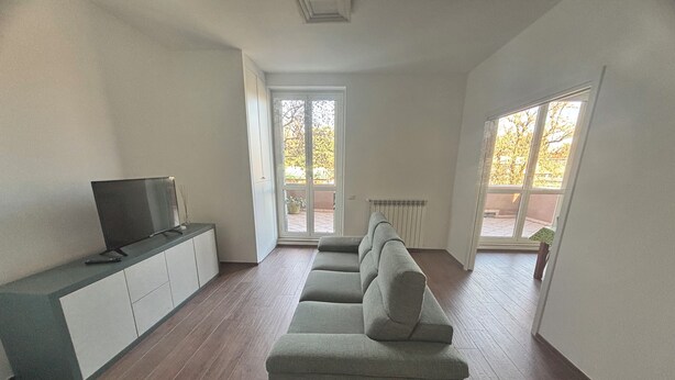 Ein geräumiges Wohnzimmer mit grauem Sofa, einem Flachbildfernseher auf einer Kommode und großen Fenstern mit Blick ins Freie.