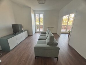 A spacious living room with a grey sofa, a flat-screen TV on a cabinet, and large windows offering a view of the outdoors. - Quiet apartment in serviced area 10 minutes' drive from Milan. (Bettola-Zeloforamagno)