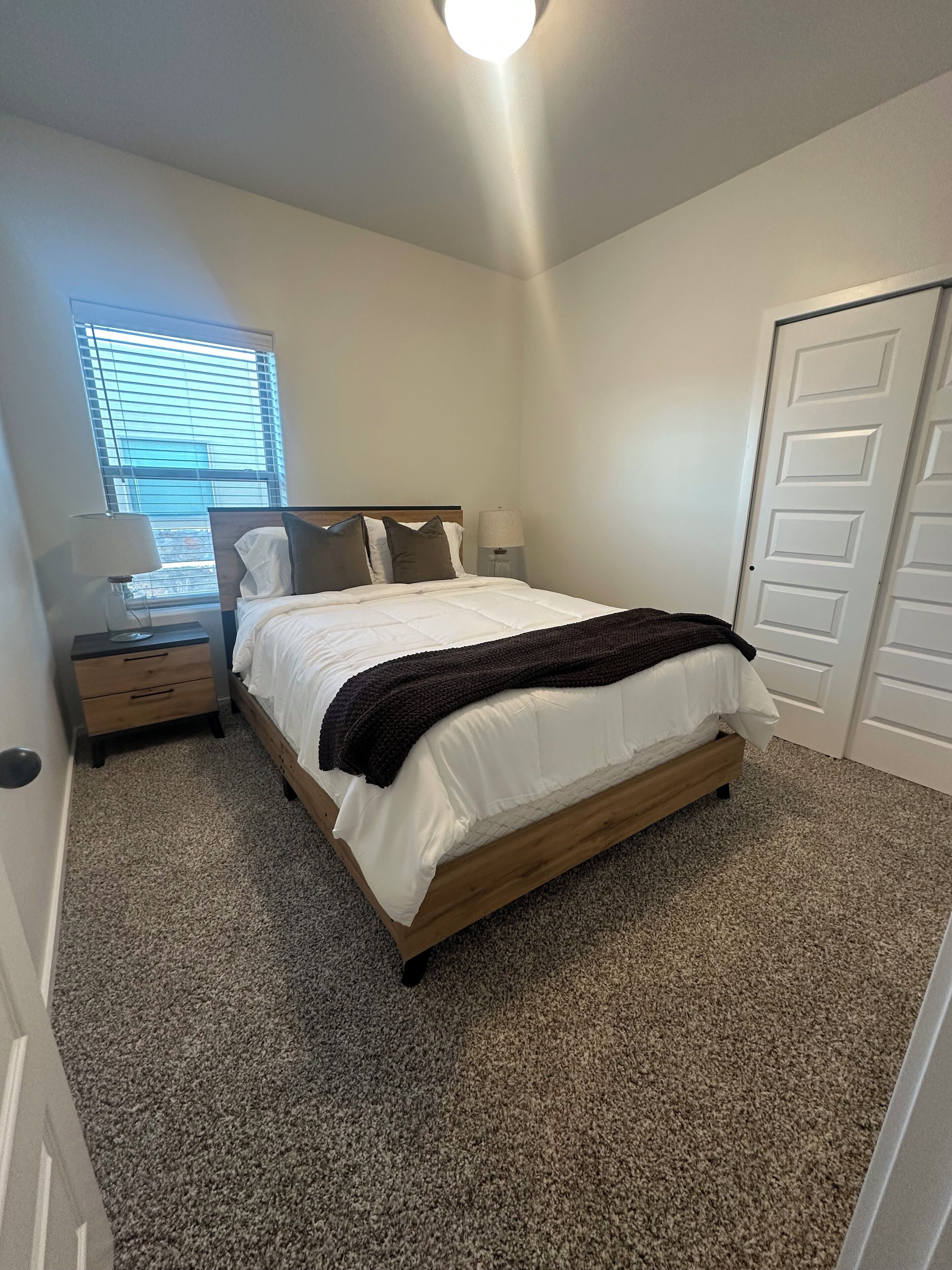 Une chambre à coucher comprenant un lit, une table de chevet, une lampe, une fenêtre avec des stores et une porte.
