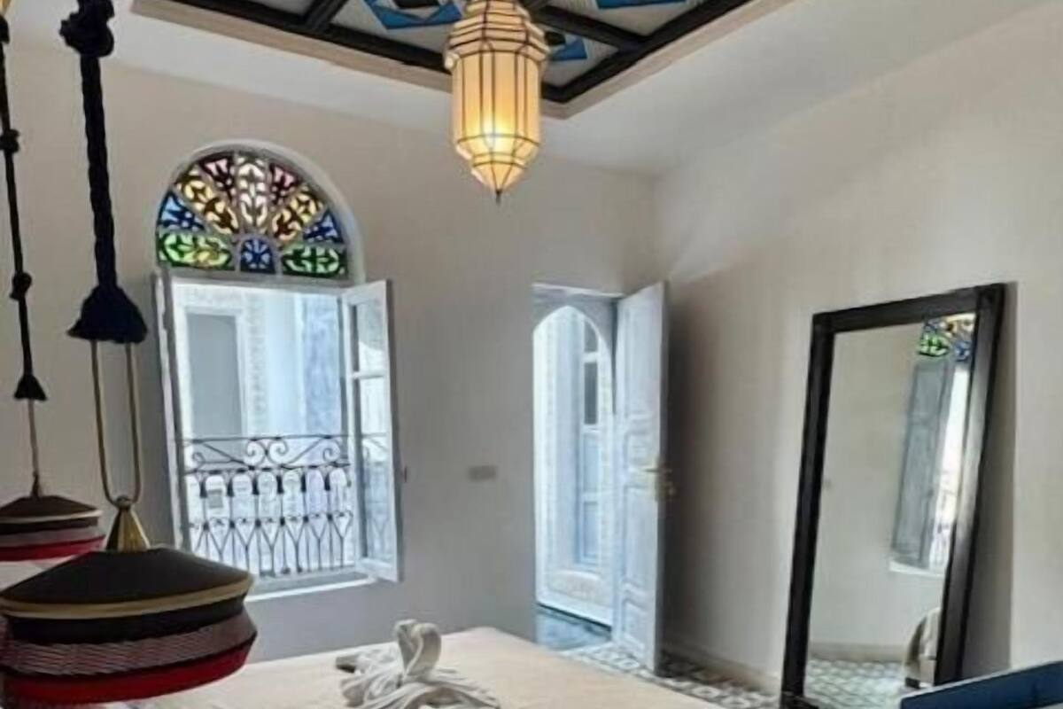 Un dormitorio con cama, lámpara colgante, ventana con celosía decorativa y espejo.