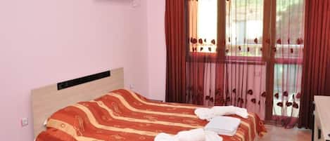 A hotel room with a bed, a nightstand, an air conditioning unit, and a window with curtains.