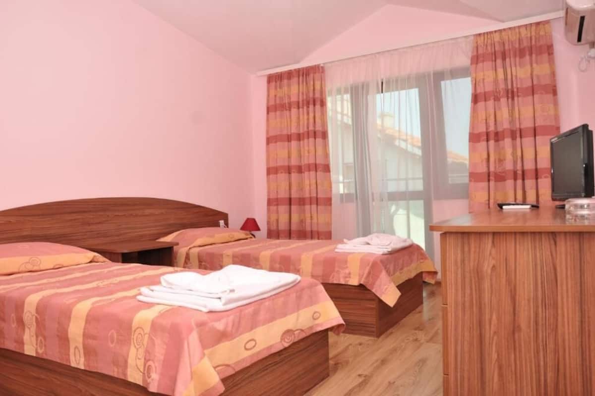 A hotel room with two beds, a wooden desk, a television, and a window with curtains.