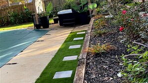 Un jardín ajardinado con un sendero pavimentado, una terraza de madera y una diversidad de plantas.