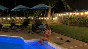 Una piscina con una persona sentada en el borde. Se ven sillones de playa y sombrillas al fondo.