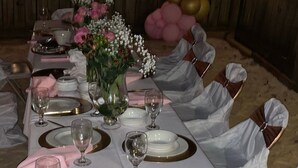 Una larga mesa de banquete dispuesta con manteles blancos, platos con borde dorado y servilletas rosas.