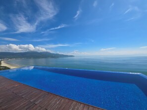Cielo azul despejado, una terraza de madera y una piscina con vistas a una playa y al océano.
