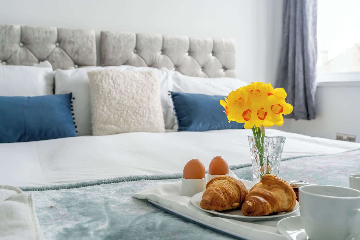 Un llit ben fet amb un edredó blanc i blau, un panell de capçal grís amb guarniments, i un ram de flors grogues sobre un plat amb croissants i ous.