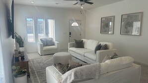 Un salon avec un canapé blanc, un fauteuil blanc, un ventilateur de plafond et deux tableaux accrochés au mur.