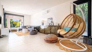 Een moderne woonkamer met een hangstoel, een kleurrijk poef en een groot raam met uitzicht op groen.