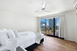 Een hotelkamer met een bed, een plafondventilator, een groot raam met gordijnen en uitzicht op een stadsgezicht.