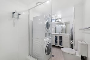 A modern bathroom with a glass shower enclosure, a stacked washer and dryer, a toilet, and a sink with a mirror. - Stylish 2BDR Retreat in Broadbeach Sleeps 4 (Gold Coast)