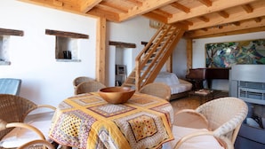 Une pièce chaleureuse avec un plafond en bois, une table ronde sur laquelle se trouve un saladier, des chaises en osier, un piano et un tableau aux couleurs vives.