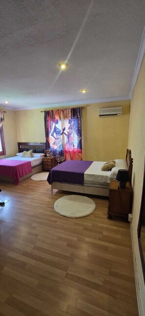A hotel room with two beds, a window with curtains, an air conditioning unit, and a wooden floor. - Villa Noa. (Torrevieja)