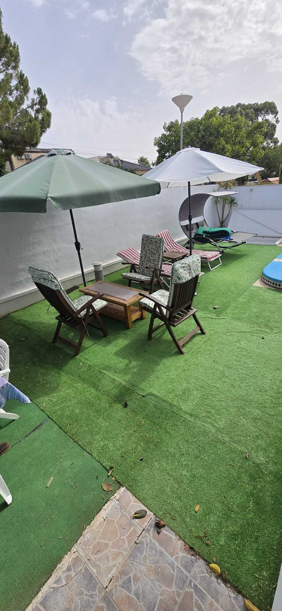 Uma área de estar externa com grama artificial, móveis de madeira e guarda-sóis.