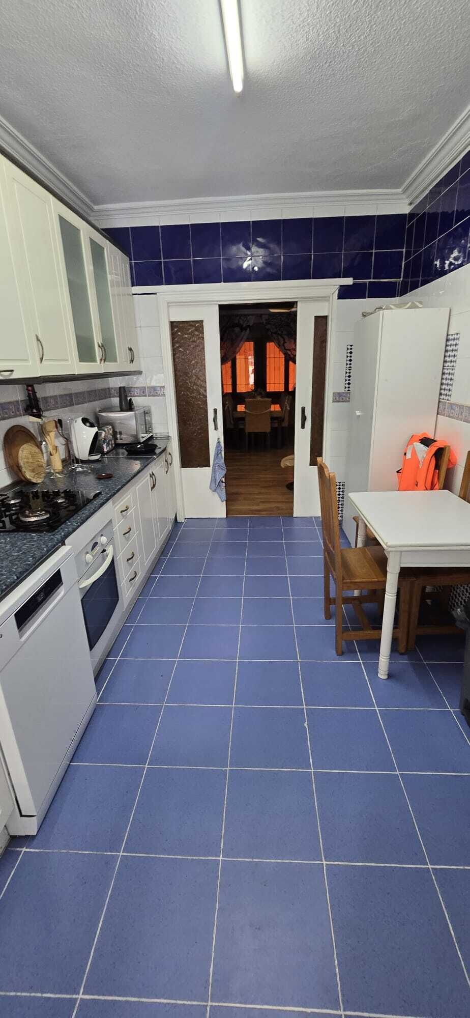 Een keuken met blauwe tegels, witte kastjes, een fornuis en een eethoek met een tafel en stoelen.
