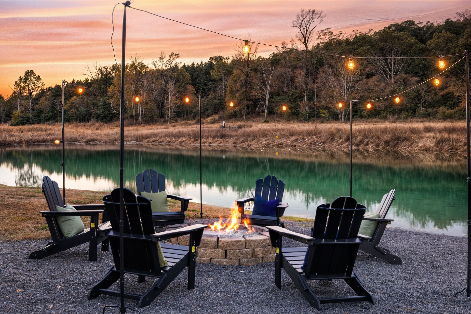 A lakeside seating area with Adirondack chairs and a fire pit, surrounded by a gravel path and string lights.