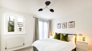 Ein Schlafzimmer mit einem großen Bett, einem Fenster mit Vorhängen, einem Heizkörper und einem Nachttisch mit Lampe und Pflanze.