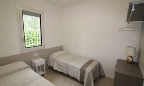 A simple, clean room with two beds, a small bedside table, a window with a view of greenery, and a lamp on a dresser. - Modern Villa With Garden in Lignano (Lignano Sabbiadoro)