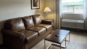 A living room with a brown leather sofa, a small table, a lamp, and a window with curtains. - Four Points By Sheraton Prince George (Prince George)