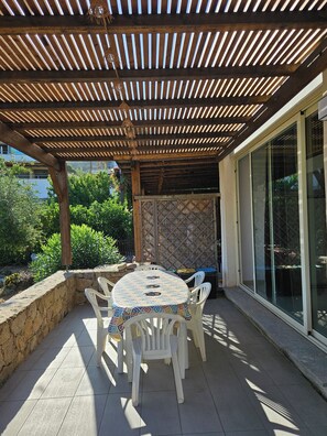 An outdoor patio with a round table and chairs, a wooden pergola overhead, and a view of greenery and a building in the background. - House in Marine di Sant'Ambroggio, 10 minutes from the ocean, large terrace (Lumio)