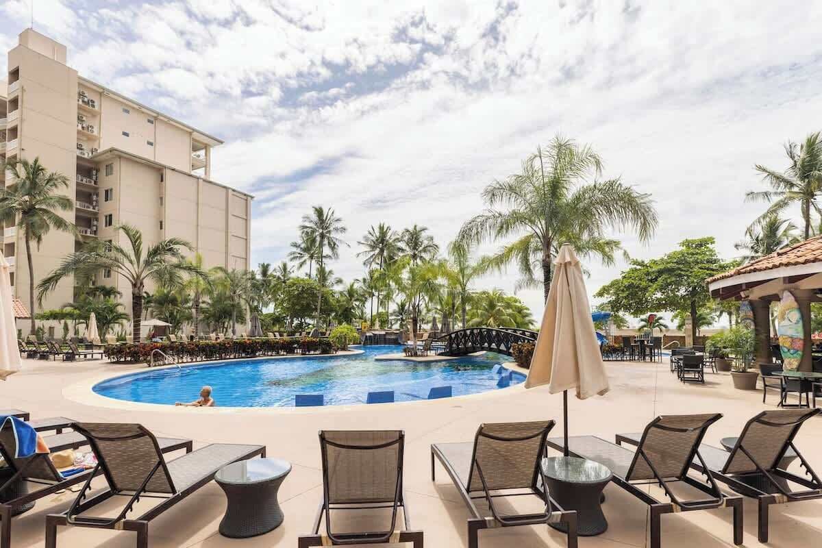 A pool area with lounge chairs and umbrellas, surrounded by palm trees and a building.