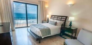 A hotel room with a bed, a nightstand, a chair, and a view of the ocean through a large sliding door.