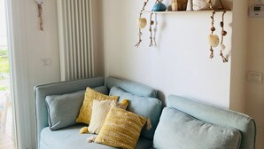 Un sofá azul claro con cojines estampados, un estante fijado a la pared con adornos, y una ventana con persianas.