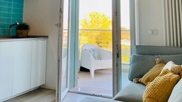 Una cocina moderna con armarios blancos, salpicadero color turquesa y puerta corrediza de vidrio que da a un balcón con una silla blanca y una manta amarilla.
