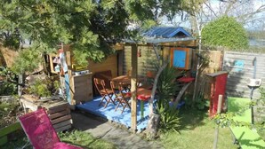 A small, rustic outdoor seating area with a wooden table and chairs, a pink lounge chair, and a small shed with a blue roof.