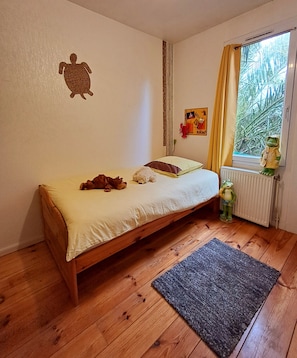 A small, cozy bedroom with a single bed, a wooden floor, a radiator, and a window with curtains. - Colorful little house (Plérin)