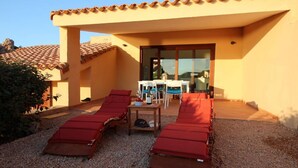 Uma área externa ensolarada com assentos ao ar livre, cadeiras reclináveis vermelhas e uma mesa, um alpendre coberto e a entrada de um prédio com porta de vidro.