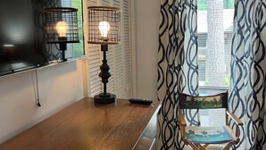 Una habitación con barra de madera, un televisor de pantalla plana en la pared, una lámpara de mesa, una silla plegable y una ventana con cortinas estampadas.
