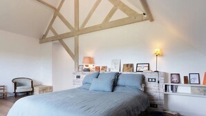Une chambre avec un grand lit, des poutres en bois, un mur en pierre servant de décor, et une étagère où sont exposés des tableaux encadrés.