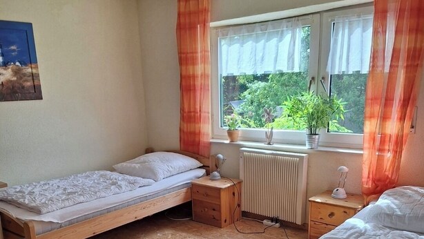 A room with two wooden beds, a window with curtains, a radiator, and a small table with a lamp. - Cozy vacation apartment at the Hainerhof (Grünberg)