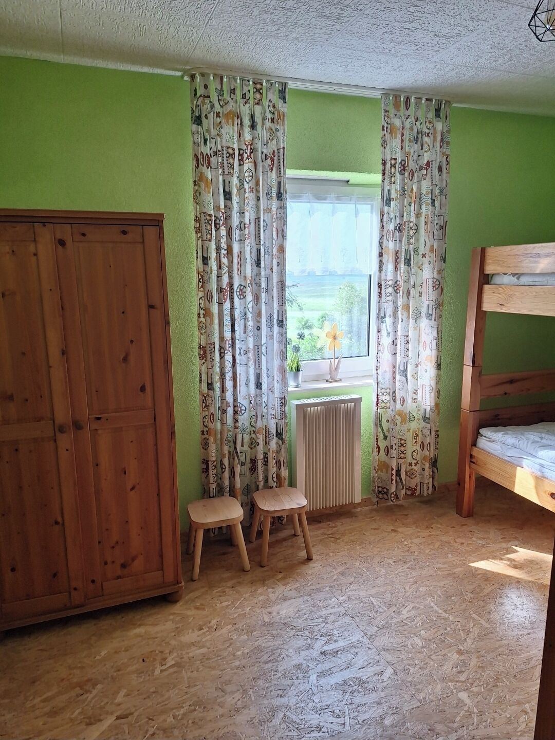 A room with a bunk bed, a wooden wardrobe, a radiator, and a window with curtains.