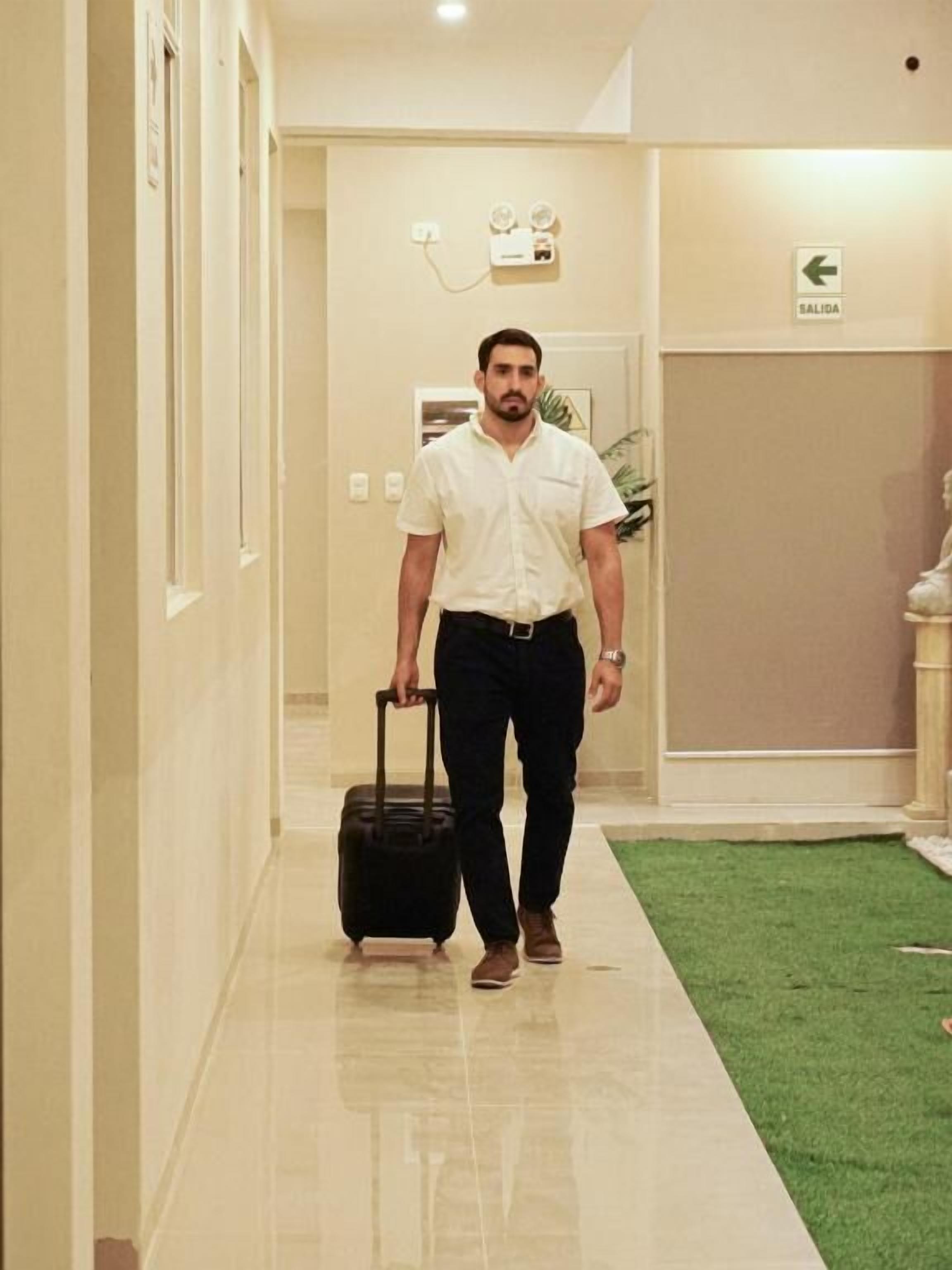 A man walking down a hallway with a suitcase, a sign indicating an exit, and a wall-mounted device.