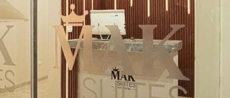 A reception area with a modern design, featuring a wooden panel wall and a glass partition. A reception desk with a computer and a sign reading "MAK SUITES" is visible.