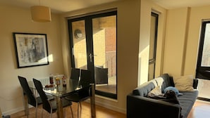Ein modernes Wohnzimmer mit einem Glastisch, schwarzen Stühlen, einer schwarzen Couch und einem großen Fenster mit Blick auf ein Backsteingebäude.