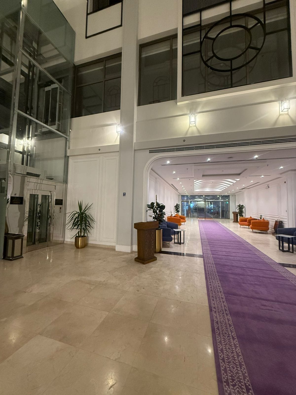 Un vestíbulo amplio con piso de mármol, alfombra morada y zonas de estar con sillas anaranjadas y azules.