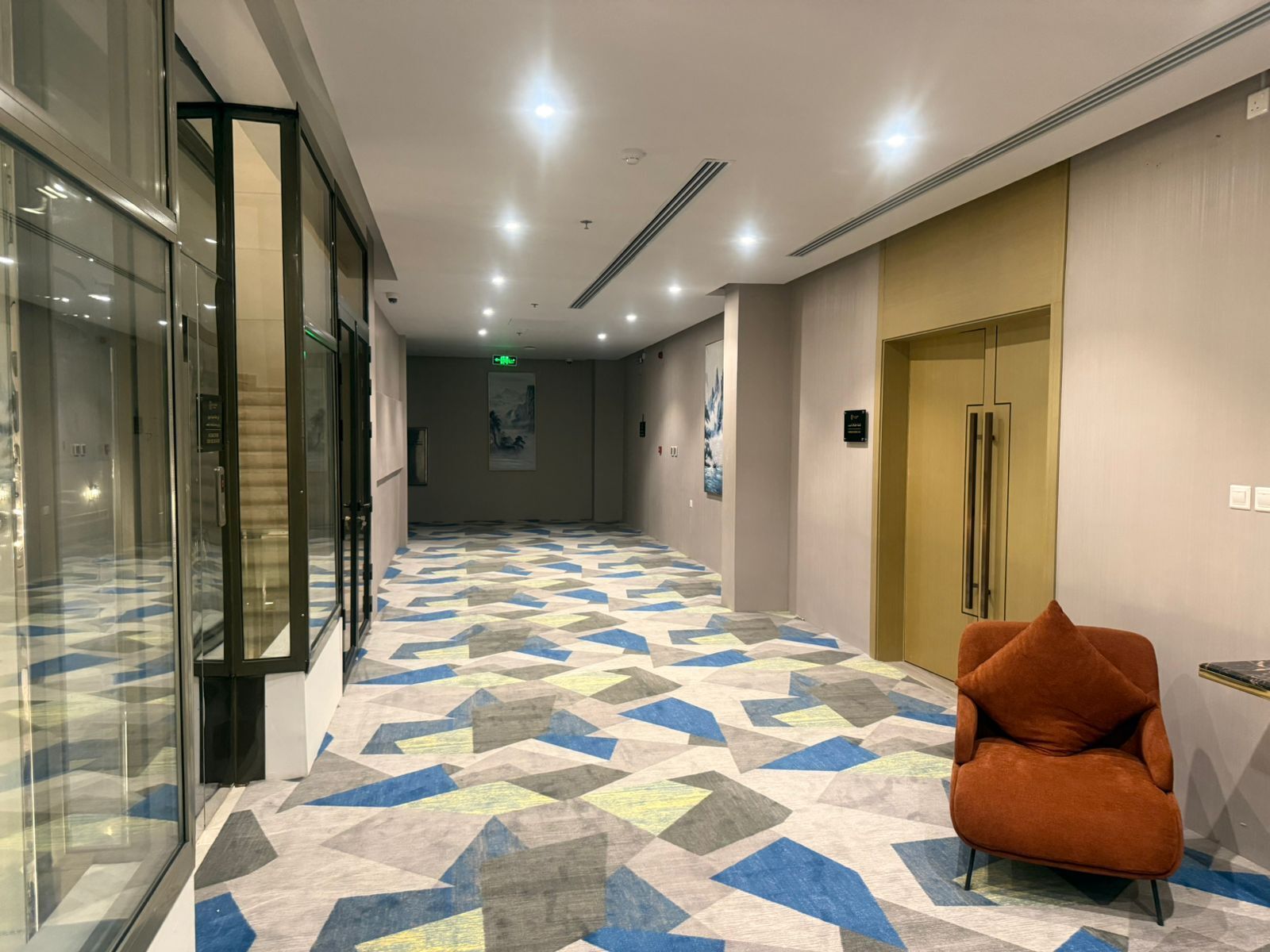 A hotel corridor with a patterned floor, a glass elevator, and a seating area with a chair.