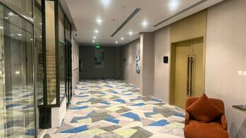 Un pasillo de hotel con piso estampado, un ascensor de vidrio y una zona de estar con una silla.
